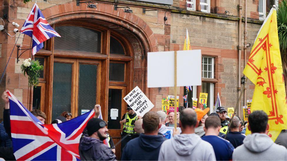 Groups of protesters outside a hotel housing asylum seekers in Perth. A Union Jack flag is being raised in the foreground as is a Lion Rampant. In the background, a protesters is holding a whote placard with the phrase 'no hate, no fear, refugees are welcome here'.