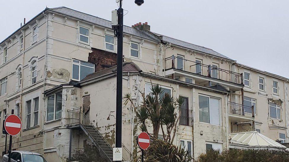 Rear view of large sprawling cream painted hotel, many of its walls have peeling paint and missing render - a road runs to the left and plants are growing along the rear.