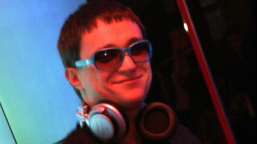 A man with close-cropped brown hair is wearing sunglasses indoors and a pair of headphones around his neck. He appears to be inside a nightclub.