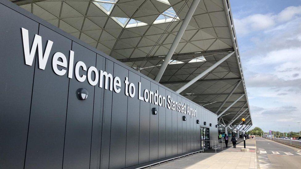 The entrance to Stansted Airport, which has a sign welcoming visitors. It has a large white roof which is supported by metal beams. The entrance is next to a road with a zebra crossing.