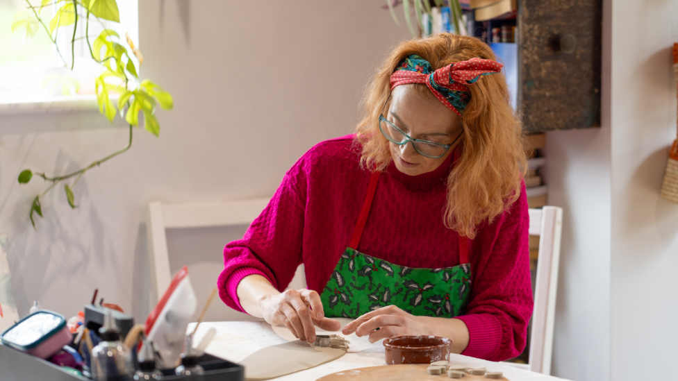 Sarah Cook is sitting at a table wearing a pink jumper and a green apron. She is using a cookie cutter to cut out hearts from a sheet of clay.