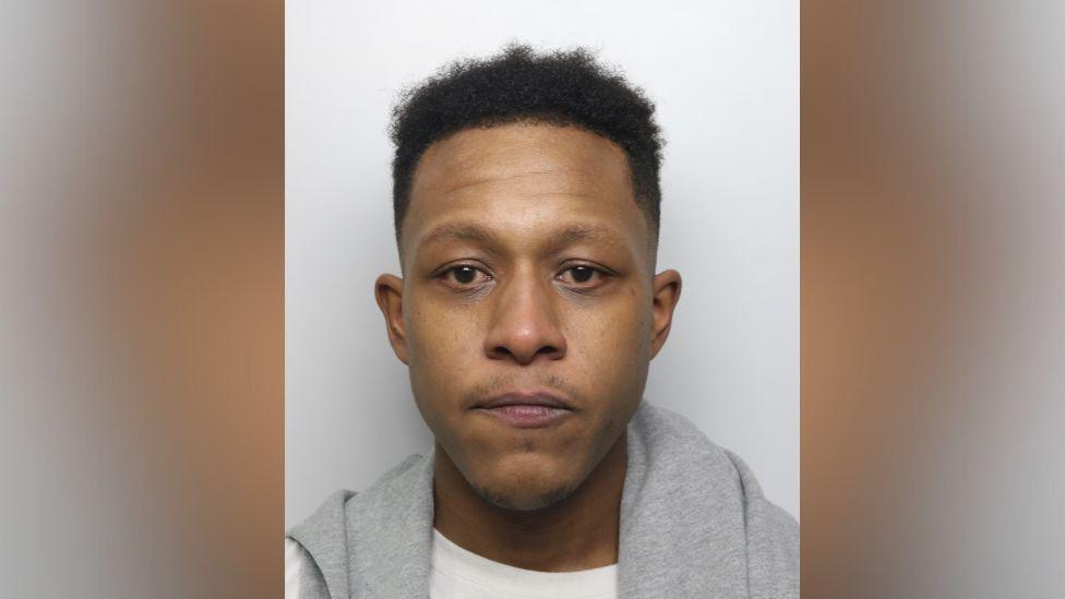 A police mugshot of Kane Clarke, a man with black hair and a grey jumper apparently draped round his shoulders. He is staring into the camera. 