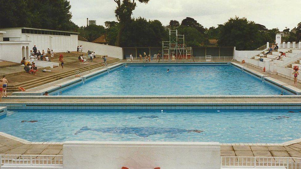 Broomhill lido pictured in 1969. Blue tiles in the shape of a whale can be seen at the bottom of the pool. There are people sitting around the edge of the pool. 