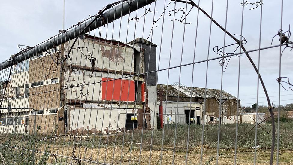 A view of a derelict factory through a silver metal fence. The building is partially demolished with broken windows and is surrounded by overgrown grass and weeds.