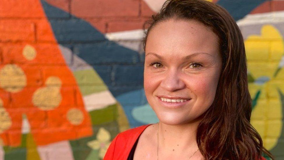 A woman with brown hair. She is standing smiling at the camera. She is stood in front of a multi-coloured mural painted on a brick wall.