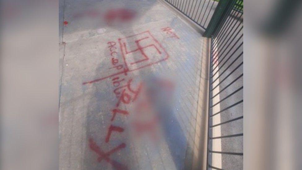 Grey ground surrounded by railings of a bandstand. You can see red spray-painted images, with a swastika symbol with a red square around it, as well as the words 'not acceptable' and 'jolly'. Part of the spray paint is blurred out.