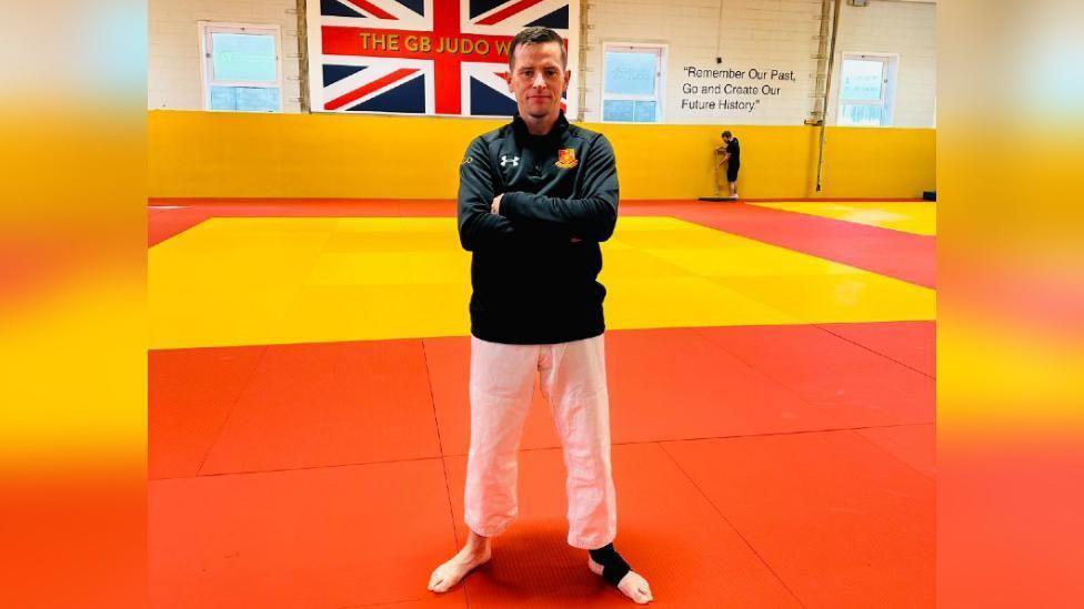 A man wearing a black jacket and white trousers, with a strap around his left foot, stands with his arms crossed in a gym, The floor is yellow and red and a union flag can be seen on the wall in the background.