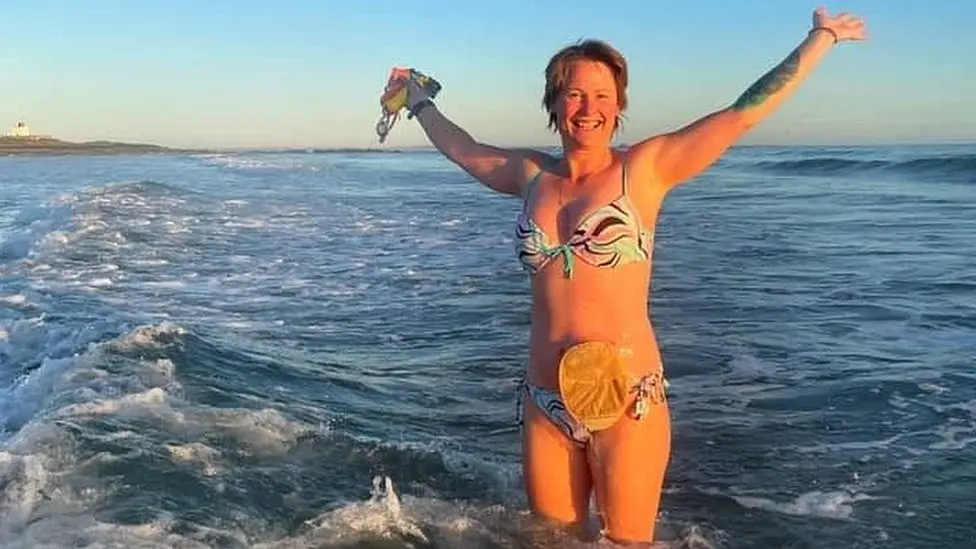 Gill Castle standing in the sea with her arms in the air. She has short, brown hair and is wearing a patterned bikini. Her stoma bag is visible to the side of her stomach.