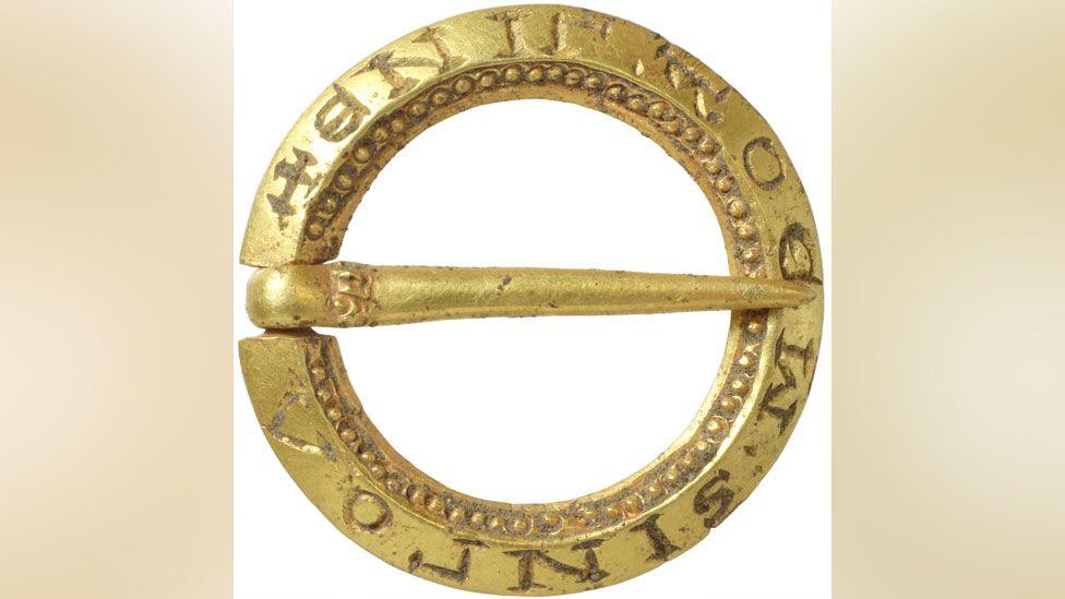 A close-up of a small, gold round medieval brooch. It is made from a ring with a gold pin crossing over its middle. The outer ring has words etched into it and the inner ring has a decoration of tiny gold dots