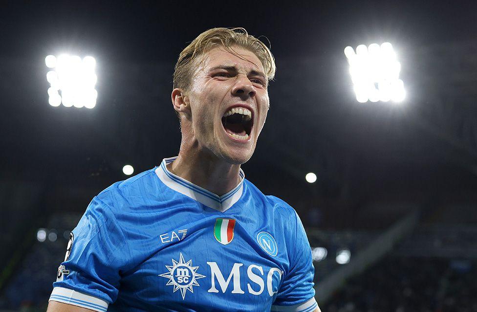 Napoli’s Rasmus Hojlund celebrates after scoring the first goal in a Champions League match against Sporting Clube de Portugal in Naples. He wears a bright blue Napoli jersey with white trim, sponsor logos, he's celebrating under stadium floodlights.