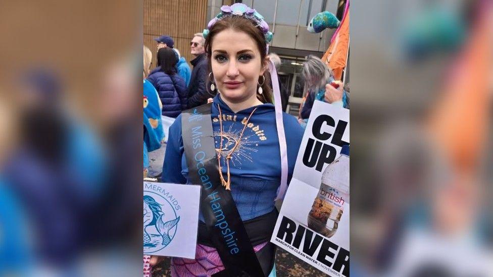 Ms Seales wearing a blue top and purple leggings, and a sash saying "Miss Ocean Hampshire". She's holding up placards calling for rivers to be cleared up.