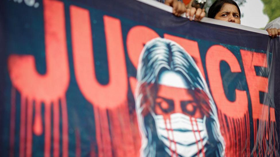 Banner with the word justice written in red, with red lines dripping from the base of the letters, like blood; an image of a woman wearing a surgical mask is also depicted