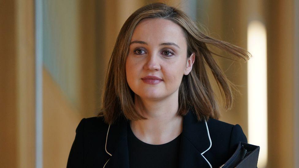 A woman with light brown hair, wearing a dark suit, in a medium close-up shot as she walks in the Scottish Parliament 