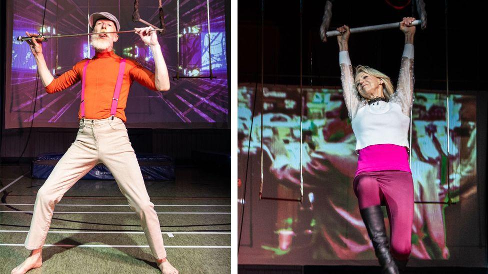 A woman swings from a trapeze by both hands, with a screen behind her showing a film. She wears a sparkly white long sleeved top with transparent sleeves, and a pink vest underneath. She wears black knee boots and shiny maroon leggings. On her left is a man holding a sword to his mouth. He wears an orange polo neck sweater, pink breeches, beige trousers and a flat cap. He has a bushy white beard.