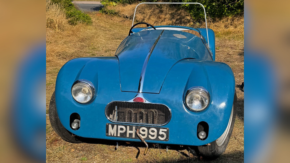 A blue Connaught L2 sports car, which has large headlights and is parked on grass. Its engine is exposed thanks to a mesh grille and it has a long bonnet. The small car has two-doors.