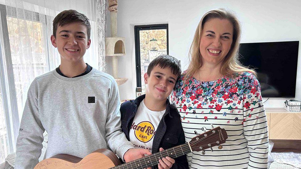Michal, Jakob and Beata Wojcik standing in their sitting room. Michael has short dark brown hair and is wearing a grey sweat shirt and holding the guitar; Jakob has short black hair and is wearing a black open shirt over a white T-shirt and is leaning his head against his mum's shoulder; Beata has fair hair falling to her shoulders and is wearing a white top with black stripes and flowers covering its top quarter. They are all smiling happily.