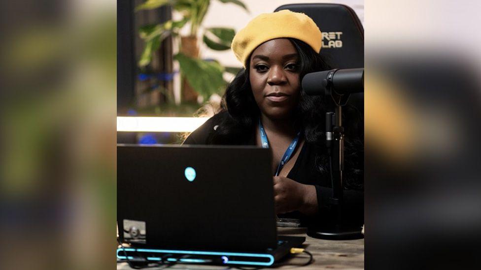 A woman in a yellow beret sits at a desk in a high-backed gaming chair. A streaming microphone is set up close to her face and there's a black laptop with a glowing alien logo in the middle of the lid open in front of her.