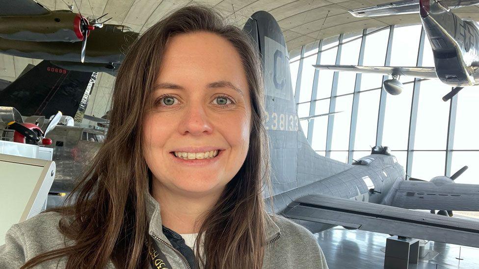 Hattie Hearn standing in the American Air Museum at the Imperial War Museum in Duxford. She has light brown hair over her shoulders and is wearing a grey sweatshirt. Behind her can be seen various aeroplanes, some suspended from the roof, and beyond that is a large window reaching up to the roof. 