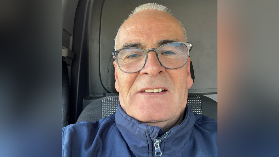 A man with short grey hair, somewhat balding. He is wearing glasses and a blue zip up jacket. He is sitting in a vehicle.