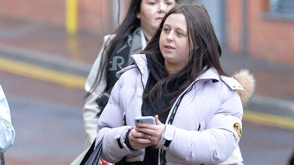 A woman with dark long hair and a lip piercing walks in a street. She is holding a phone in front of her with both hands. She is wearing a light-purple puffy jacket and holding a bag with her right arm. 