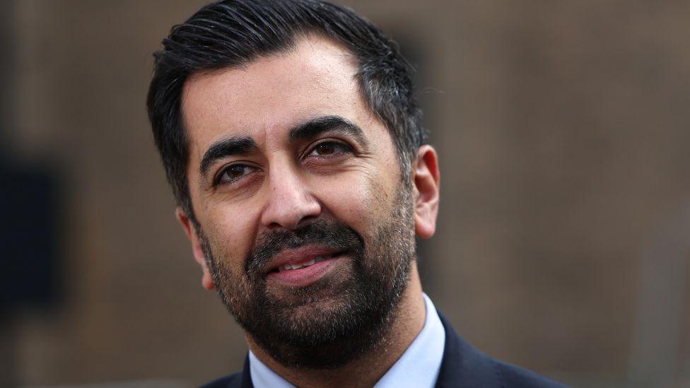 Humza Yousaf, with dark hair and dark beard, in front of a blurred brown background 