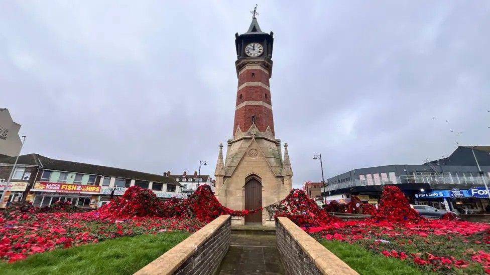 A clock tower in the middle of a roundabout has poppies either side of it. There is a grass front with a fish and chip shop in the background.