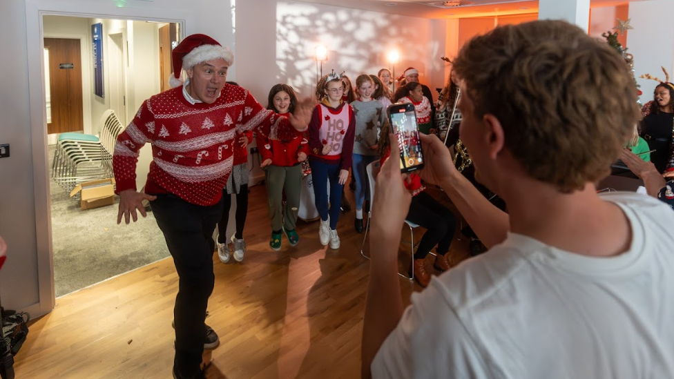 Ed Davey, wearing a Christmas jumper and Santa hat, making a funny face at someone taking a picture on their phone.