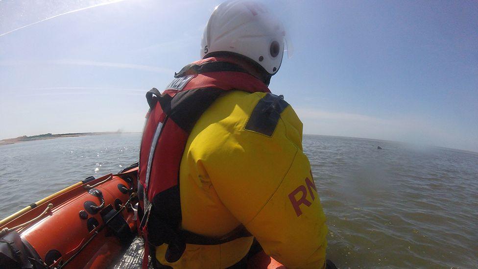 The back of an RNLI crew member in a yellow jacket and white helmet, in a small orange boat. He is looking out at the water where a casualty can be seen in the distance