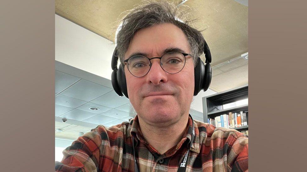  A selfie by Prof Andrew Williams, who is wearing headphones, black-rimmed glasses and a red, white and black plaid shirt. Behind him is a book shelf. 
