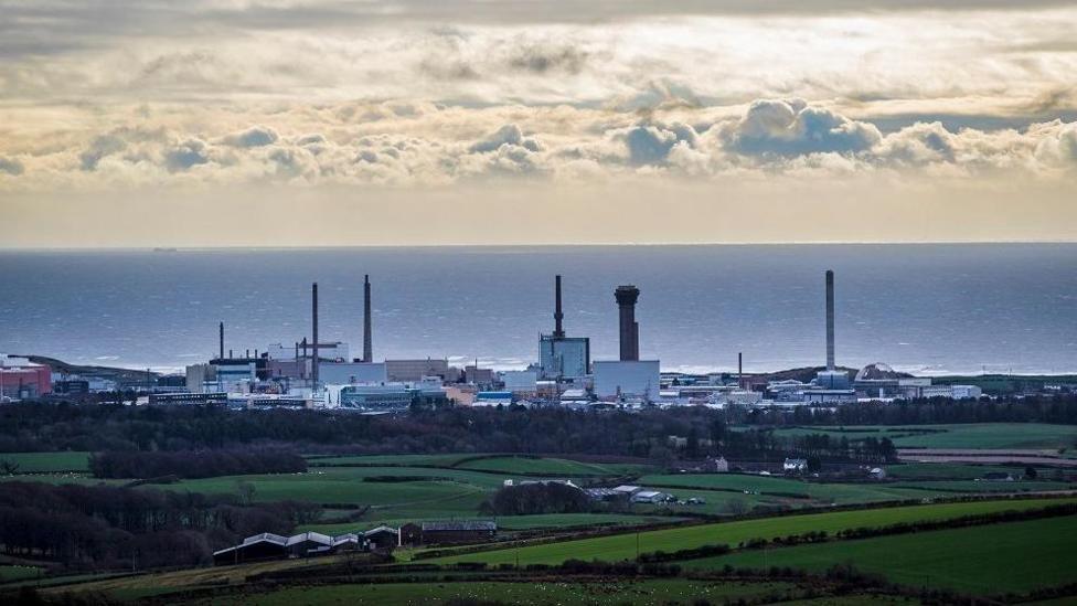 General view of Sellafield Nuclear power plant, in Cumbria. The shot is taken from a distance further away, with the sea on the horizon and a cloudy, but sunny day. The foreground is green areas of land.