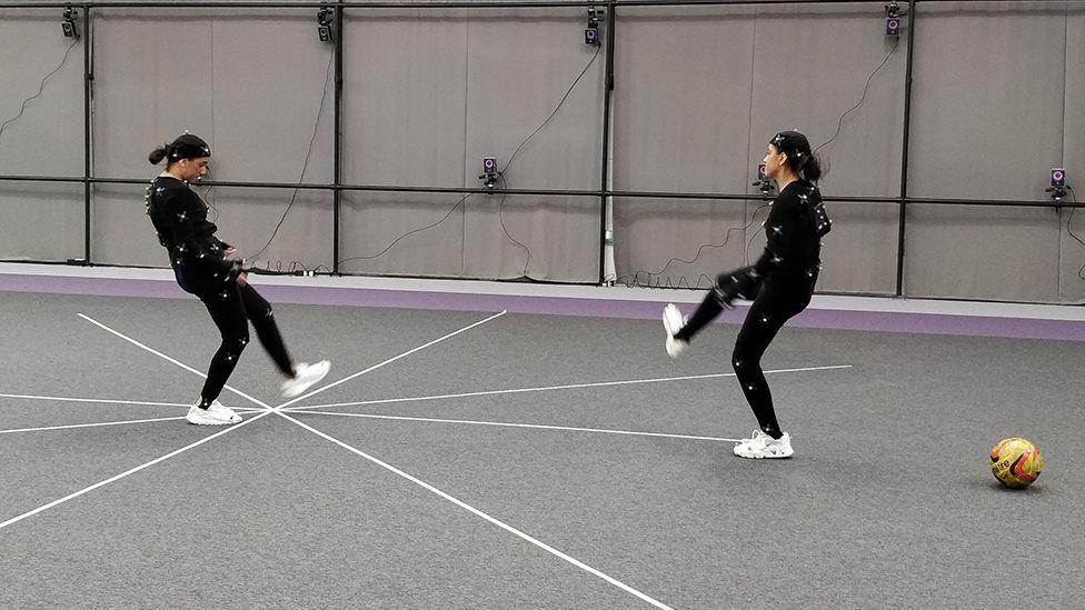 Two women wearing black, skin-tight motion capture suits with white, ping-pong ball-style spheres attached to them, make kicking motions like those you'd see in a game of football. Numerous small cameras are attached to the grey walls of the studio to record their movements from different angles.