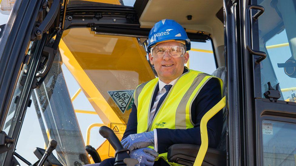 James Timpson sitting in the cab of a construction vehicle. He is wearing a blue hard hat and a yellow high-vis jacket over a navy suit. He is wearing clear  protective goggles. 