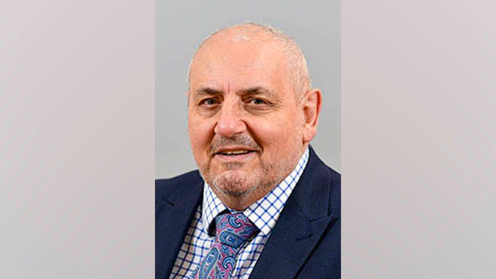 A headshot of Mark Fryer. He is middle-aged and balding with stubble on his chain. He is wearing a smart blue suit and a white shirt, with a blue criss-cross pattern. He tie has a colourful pattern of pinks and blues.