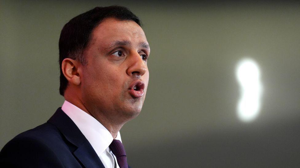 A man with dark hair, wearing a dark suit and white shirt with purple tie, photographed from a side-on angle while he speaks.