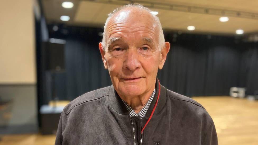 A man with short white hair, wearing a brown jacket