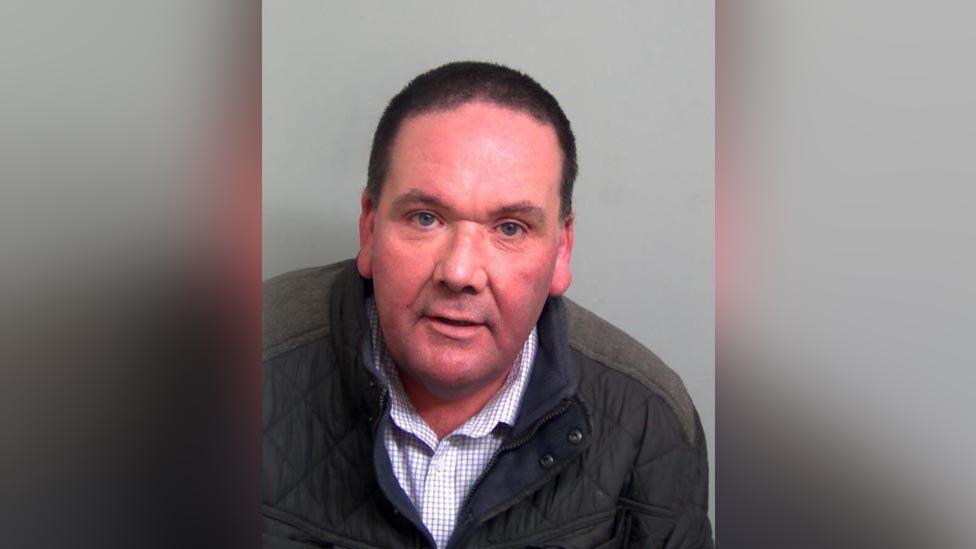 A custody photo of Craig Sharp. He is facing the camera with his mouth slightly open. He has short, dark hair. He is wearing a dark green jacket with a blue and white chequered shirt underneath.