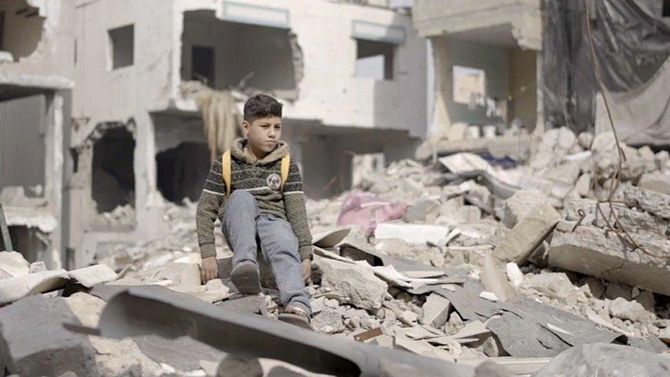 Tareq, a 10-year-old Palestinian boy, sits on a pile of rubble in Gaza, wearing jeans, a sweater, and a yellow backpack