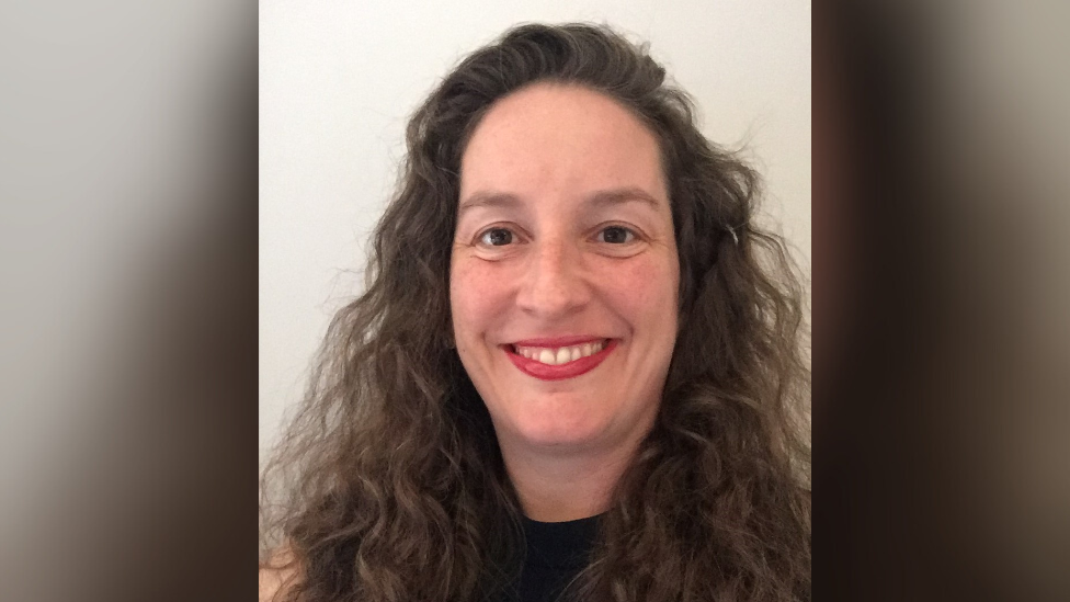 Prof Rachel Isba is smiling at the camera while taking a photo of herself against a white background. She has long curly brown hair, brown eyes and is wearing a black top and red lipstick. 