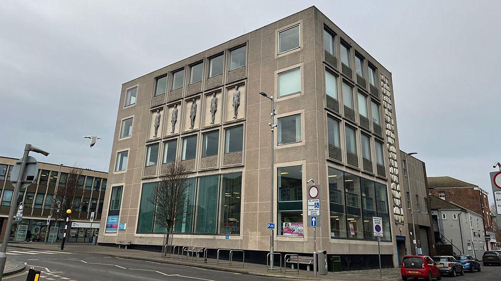 The exterior of Grimsby Central Library. It is a five-storey brutalist concrete building with double sized windows to the ground and first floor and decorated with sculptures of people on the fourth floor and a stone mosaic on one side 