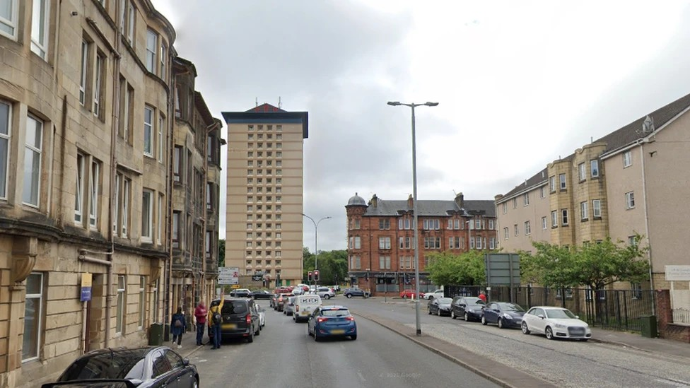A street view of Maxwellton Street street in Paisley, with blocks of tenements and more modern flats either side of a wide road with cars parked on both sides.