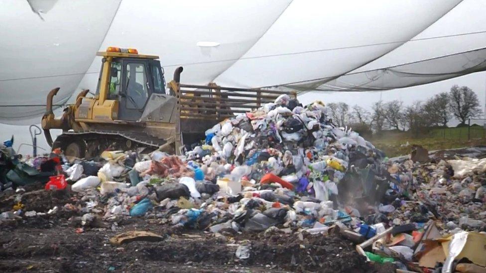 A yellow digger pushes lots of waste around a landfill site. Hills and trees are visible in the background. 