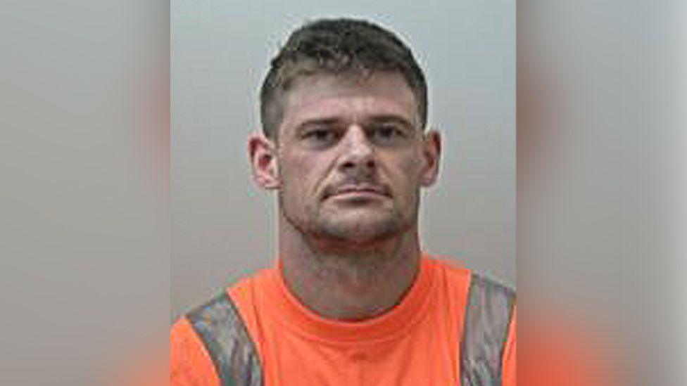 Police custody image of Adam Garnett with short brown hair and orange t-shirt with a grey strip down the shoulders.