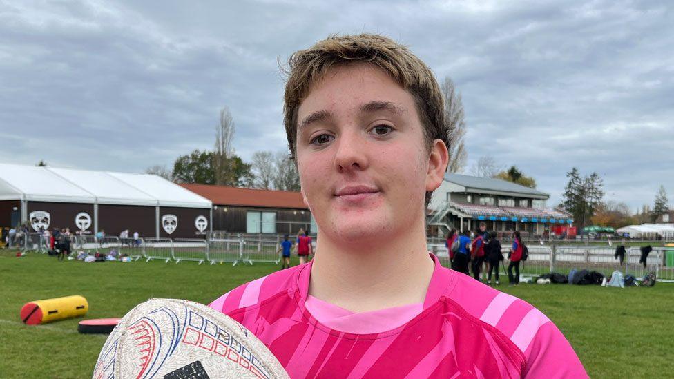 A mid-teenage girl with short hair and wearing a pink rugby shirt and holding a white rugby ball. She is standing on a pitch and behind her can be seen club buildings.