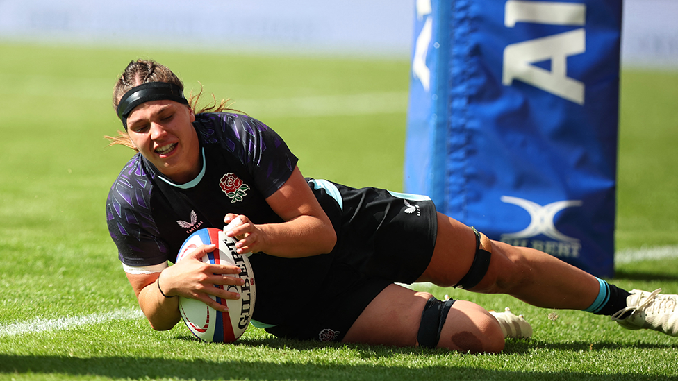 Lilli Ives Campion scoring a try for England. She is wearing the team's blue kit with a black headband and is sliding on her side to ground the ball