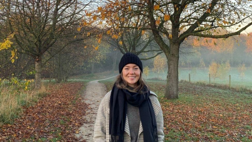 A woman with brown hair in a black beanie and black scarf stands in an autumnal landscape with fog, trees and brown leaves