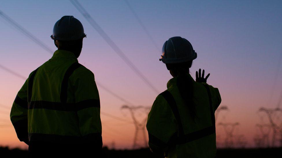 A silhouette of two engineers in hardhats at sunset with their backs to the camera looking at lots of pylons in South Africa. The woman holds up her hand towards them.