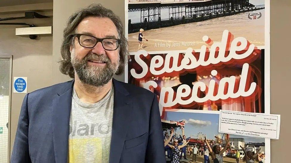 Jens Meurer wearing a navy blazer stood against a poster with pictures of Cromer beach and pier with the words, Seaside Special, a film by Jens Meurer.