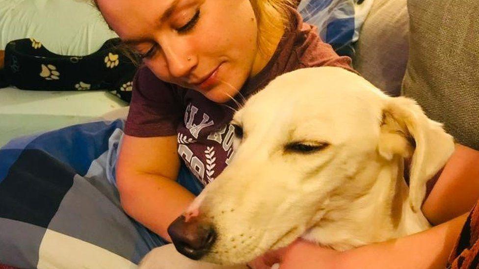 Anna Forrester is sitting on a sofa with a large light-coloured dog. She is wearing a T-shirt and is cuddling the animal