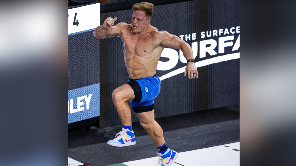 Noah Ohlsen running in the trainers painted by Ms DeLuna at a CrossFit event. He is topless, and is wearing blue shorts and socks and white Nike trainers with a blue, red and yellow design. The background is black with white writing.