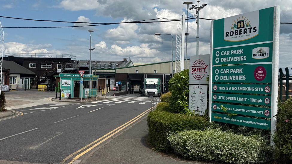 The entry to Roberts Bakery, where a green sign with directions to the car park and deliveries stands in front of a gate and a green security hut. In the background, a green and dark brown shed stands with a white Roberts-liveried lorry moving out of it and other factory buildings can be seen
